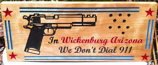 Wooden sign with a gun engraving and text 'In Wickenburg Arizona We Don't Dial 911' on an American flag background.