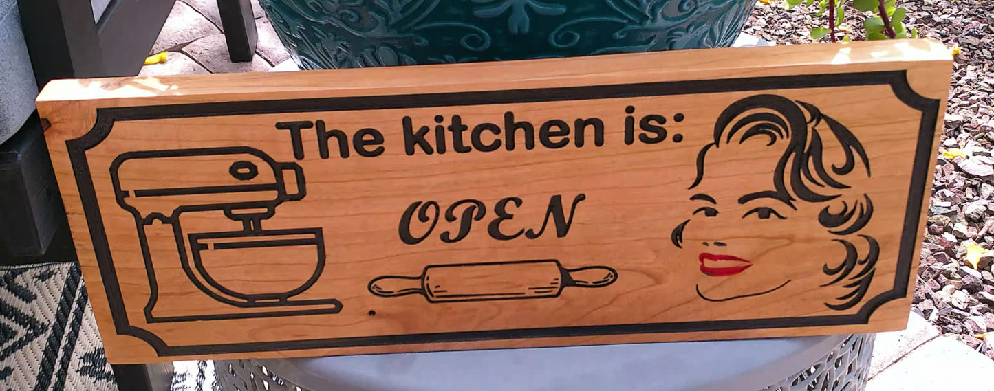 Wooden sign with a mixer, woman's face, and rolling pin design, indicating 'The kitchen is open'.