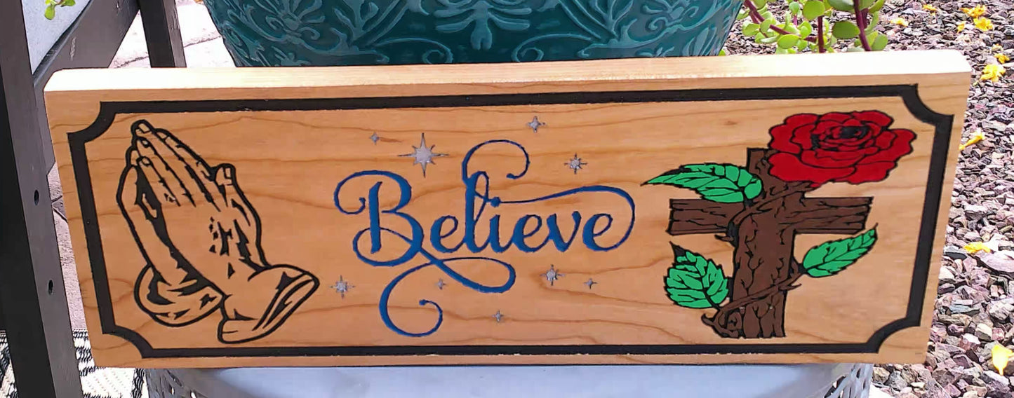 Wooden sign with praying hands, 'Believe' text, and a rose on a natural background
