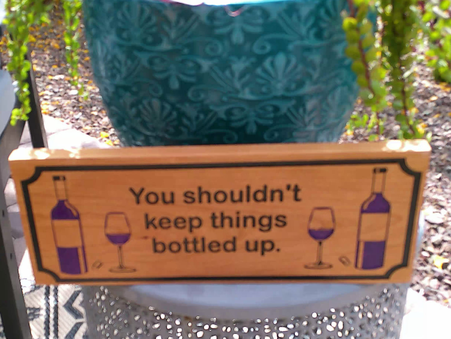 Wooden sign with humorous message and wine graphics in front of a teal cushion.